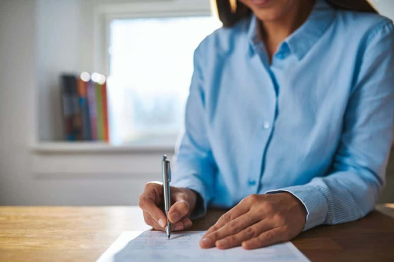 Mulher de camisa social escrevendo com uma caneta em uma folha de papel, sobre uma mesa de madeira. É fácil estudar redação