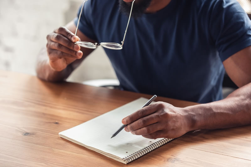 Homem inclinando-se em direção ao caderno, com um lápis em uma mão e óculos na outra.