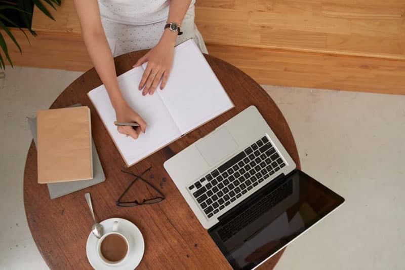 Braços femininos escrevendo em um caderno sobre uma mesa, que contém um laptop, óculos, cadernos e café