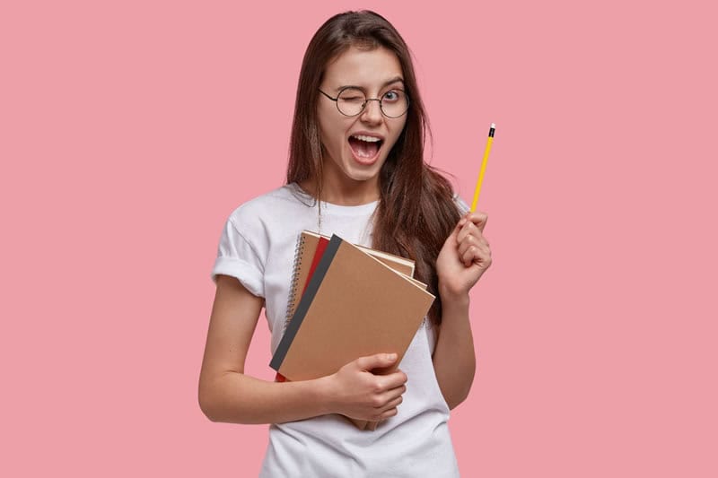 Menina segurando livros e lápis e piscando o olho, contente.