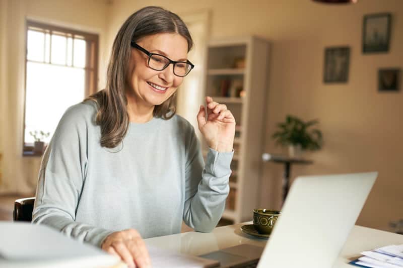 Professora de meia idade usando óculos, sorrindo e digitando prompts chatGPT em um notebook